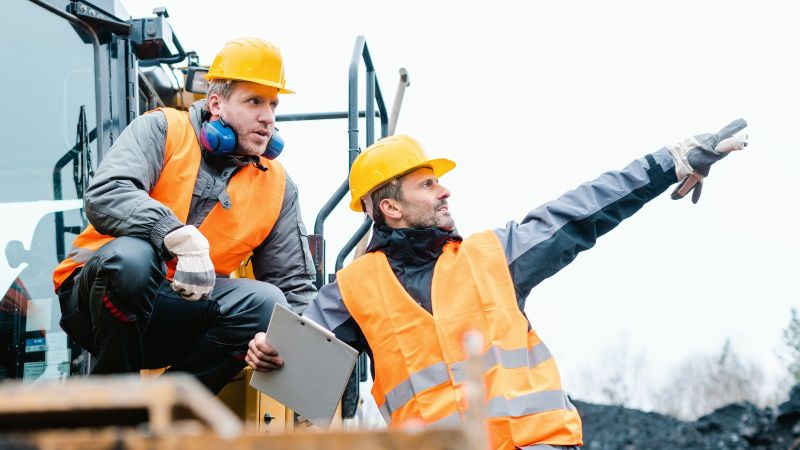 Zwei Männer (Bauarbeiter) in orangfarbener Schutzkleidung auf einem Fahrzeug, einer zeigt mit dem Finger weit weg.